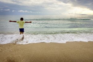 boy in a green shirt with outstretched arms facing the ocean on the beach-a mother's love