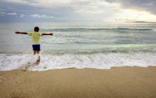 boy in a green shirt with outstretched arms facing the ocean on the beach-a mother's love