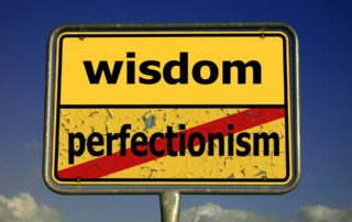 yellow road sign with the word wisdom and below it the word perfectionism marked out with a red line-perfect is the enemy