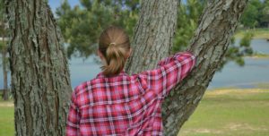girl wearing a pink flannel shirt leaning against a tree overlooking a lake