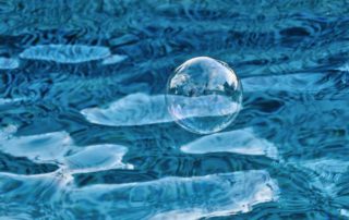 A shimmering soap bubble floats gracefully above the rippling surface of a blue pool, reflecting light and surrounding patterns.