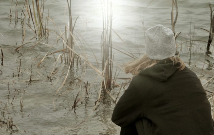 girl facing the lake amid the cattails with a burst of sun reflecting on the water as she sits alone