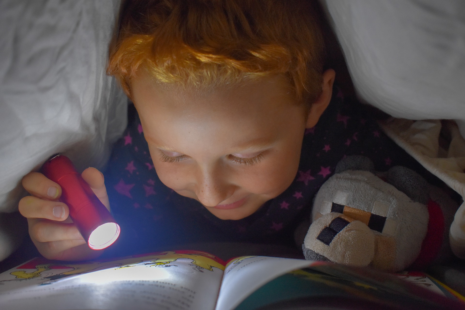 red-headed boy hiding under the covers with a flashlight learning and reading a book with his stuffed animal