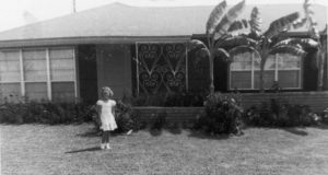 black and white photo of young girl standing in front of a 1950s house in the suburbs-houses bring memories