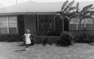 black and white photo of young girl standing in front of a 1950s house in the suburbs-houses bring memories