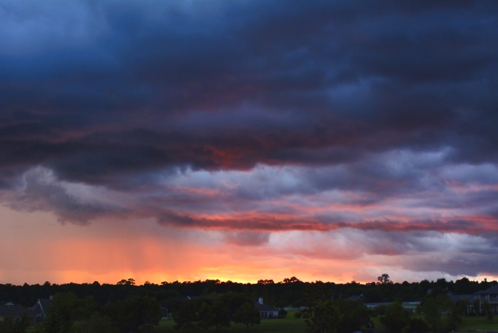 Dramatic sunset with vibrant orange and yellow hues, silhouetted trees on the horizon, and dark, billowing clouds creating a powerful, moody atmosphere.