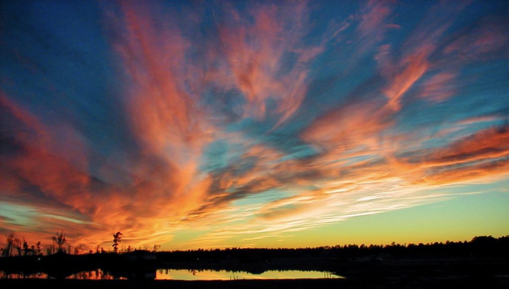 Vibrant sunset with fiery orange and pink clouds spread across a deep blue sky, reflected in a calm lake with silhouetted trees lining the horizon.
