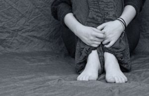 black and white photo of a person sitting only showing their crossed hands and bare feet signifying the invisibility of the victims of human trafficking