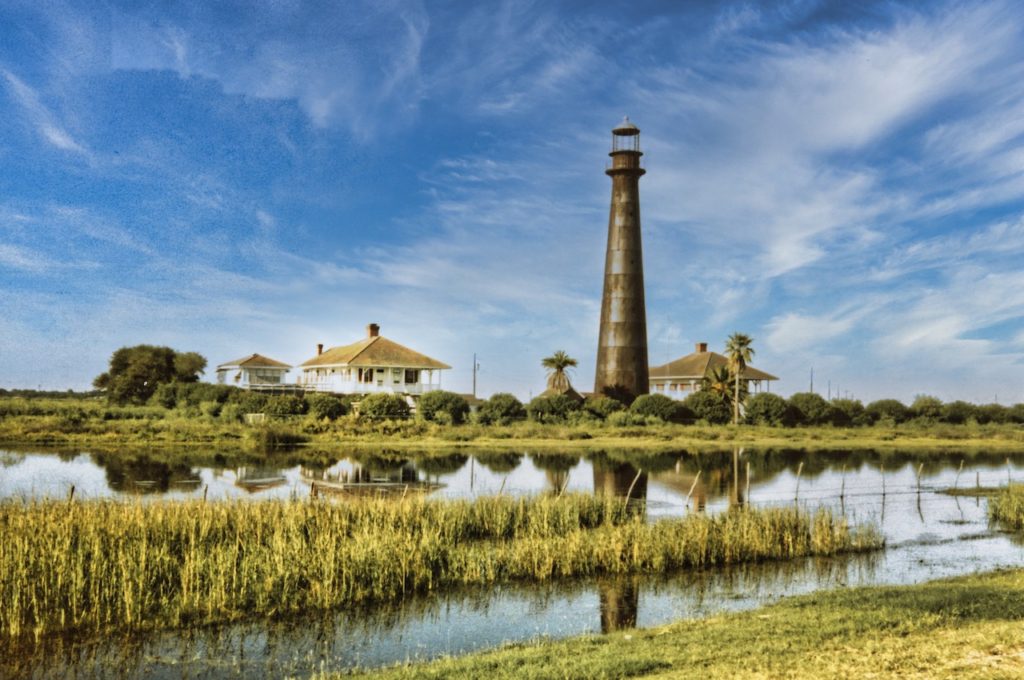 The lighthouse on Boliver peninsula stands behind the marsh surrounded by thee wooden houses on stilts with a blue sky