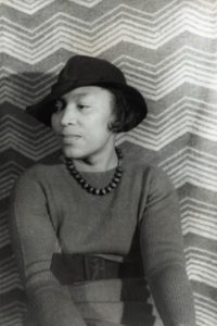 black and white portrait of Zora Neale Hurston, 1938, wearing black hat, dark belted dress and looking toward the left