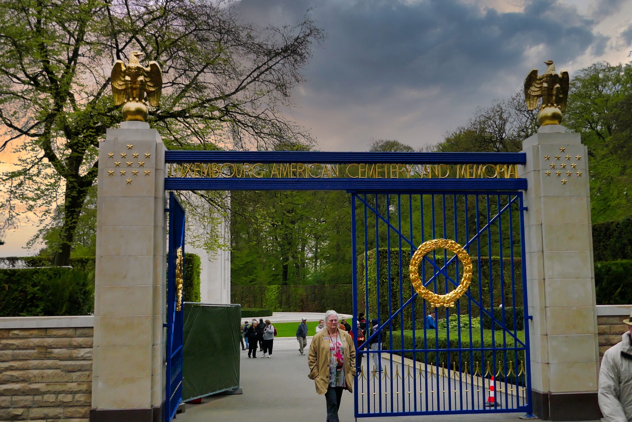 opened blue iron gate with gold wreath at the Luxembourg American Cemetery. A cloudy sky frames this door of remembrance