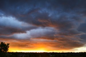 dark black and grey clouds that turn orange to gold as it meets the horizon with dark outlines of trees and houses framing the bottom of the photo-reasons to hold onto hope