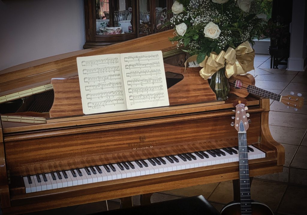tiger wood baby grand piano with sheet music, a guitar, ukulele, and white roses in a vase 