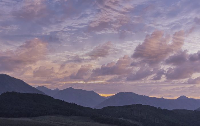 sunrise over the mountains with pink, blue, and gray clouds with a hint of the rising sun at the peak of the mountains
