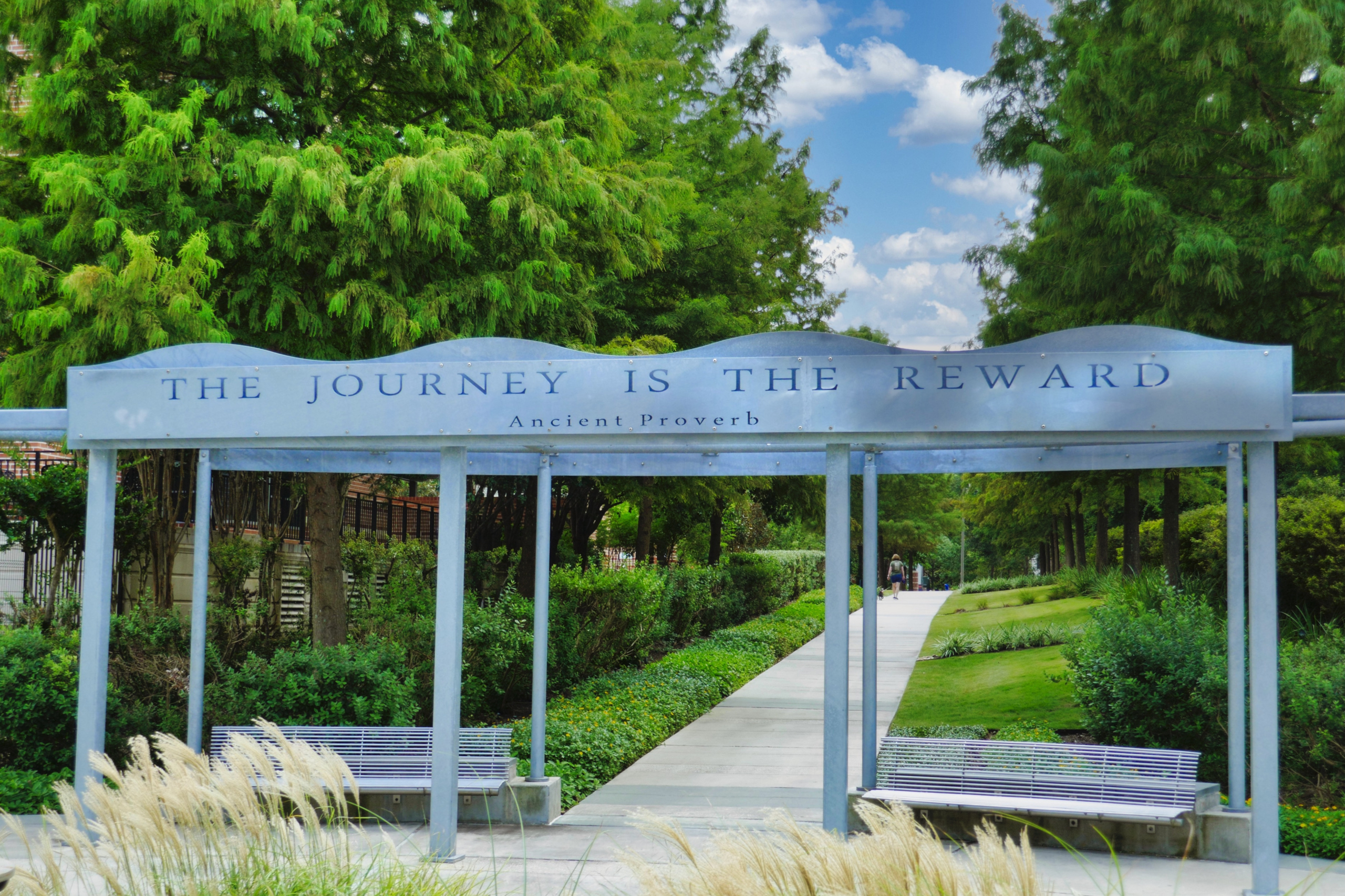 Two benches divided by a sidewalk are covered by a pergola with this ancient proverb written on the top board, "The Journey Is the Reward." Grasses sway in the foreground and green grass and trees line the sidewalk; the blue sky and puffy white clouds peek between the trees on either side of the walkway