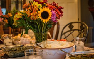 snapshot of a portion of a dinner table with a bouquet of flowers in the center, a bowl of mashed potatoes a pie to the right and a few water glasses