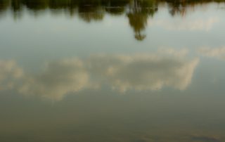 a reflection in the water of the blue sky, white clouds, and trees along the lake shoret