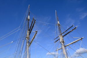 Masts and rigging of a sailing ship against a clear blue sky with a sliver of moon barely visible ibehind the intricate lines and structure.