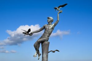 A bronze sculpture of a figure seated on a pedestal, reaching out to birds flying overhead against a bright blue sky.
