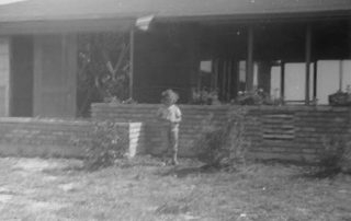 A blurred black-and-white photo of a child standing by a brick wall in front of a house with a partially visible porch.