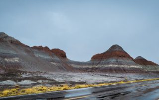 The Painted Desert Colorful striped rock formations rise beside a wet road under a cloudy sky, showcasing the beauty of a unique geological landscape.