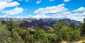 Red Rock State Park Vast landscape featuring towering red rock formations, scattered greenery, and a bright blue sky with fluffy clouds, hinting at distant snow-capped peaks.
