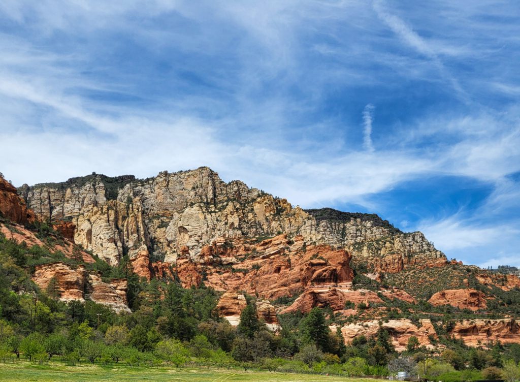 Slide Rock State Park Dramatic red and beige rock formations rise against a blue sky streaked with wispy clouds, surrounded by lush green trees.