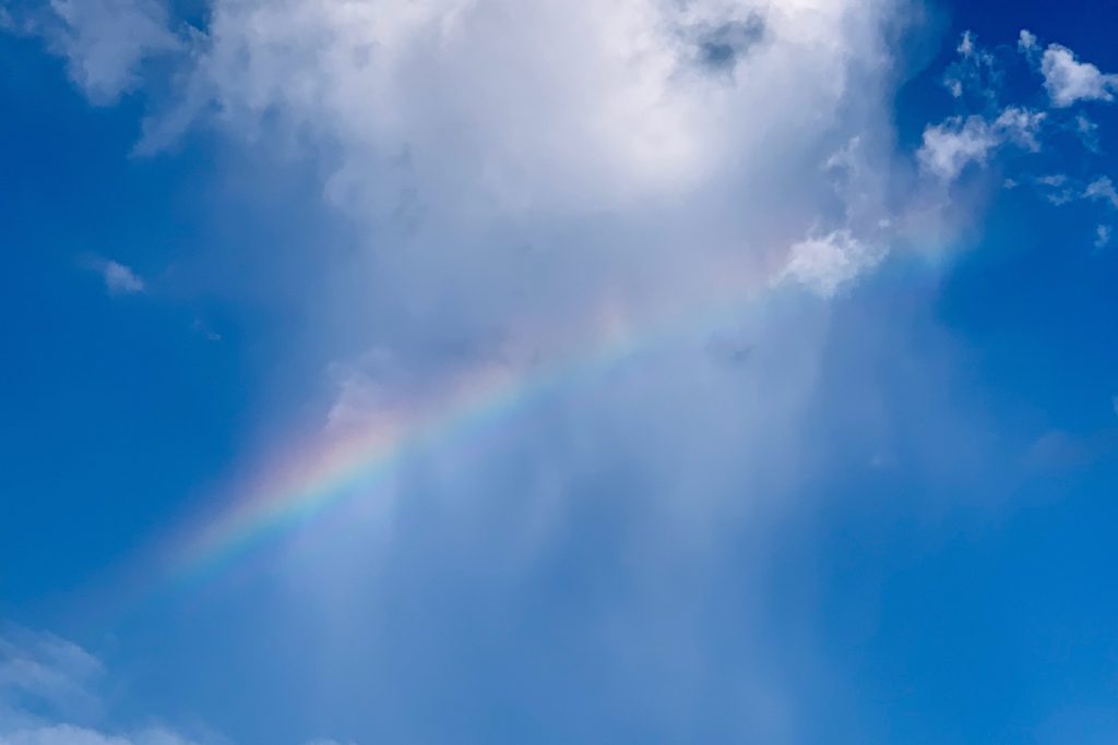 A soft rainbow arches across a bright blue sky, partially obscured by wispy white clouds.
