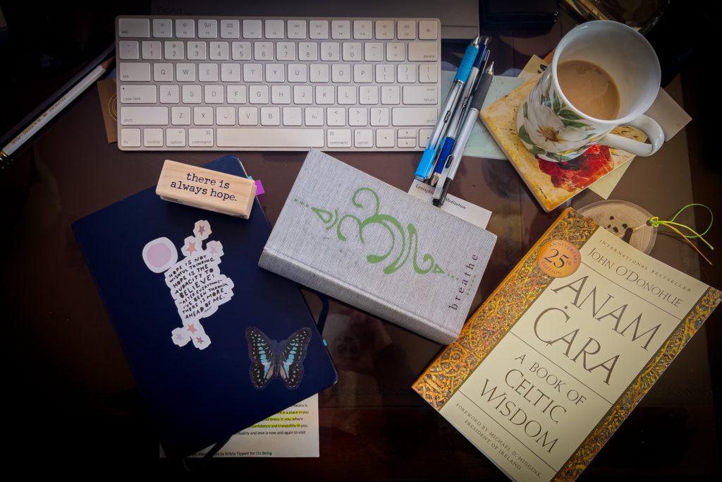 At Home © Kathryn LeRoy A workspace featuring a keyboard, notebooks, pens, a coffee cup, and a wooden block inscribed 'there is always hope.'