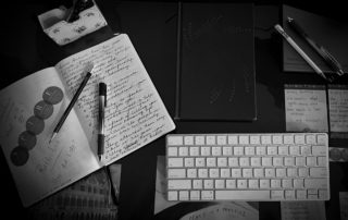 A black-and-white desk scene featuring notebooks, pens, a keyboard, and handwritten notes about reflection and personal growth.