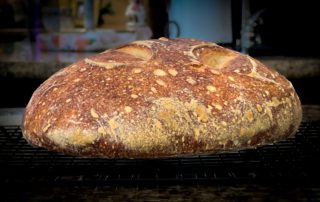 A freshly baked round loaf of artisan bread sits on a cooling rack, showcasing a crusty, golden-brown exterior and airy texture.