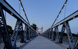 A pedestrian suspension bridge at dusk, lined with lights, leading toward a central arch structure against a deepening blue sky.