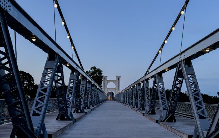 A pedestrian suspension bridge at dusk, lined with lights, leading toward a central arch structure against a deepening blue sky.