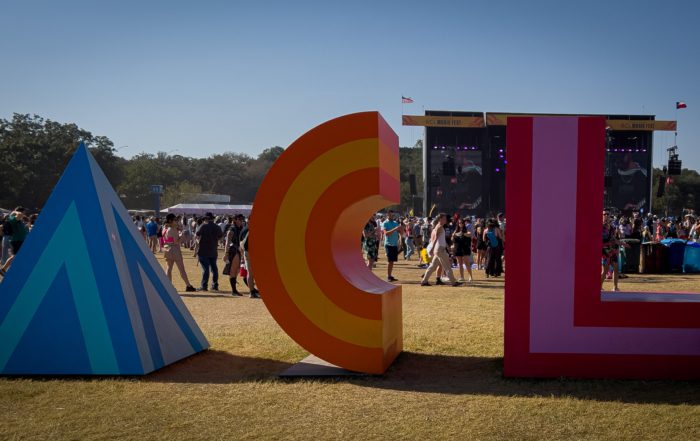 Colorful ACL festival letters stand prominently in the foreground, with a lively crowd and stage in the background under a clear blue sky.