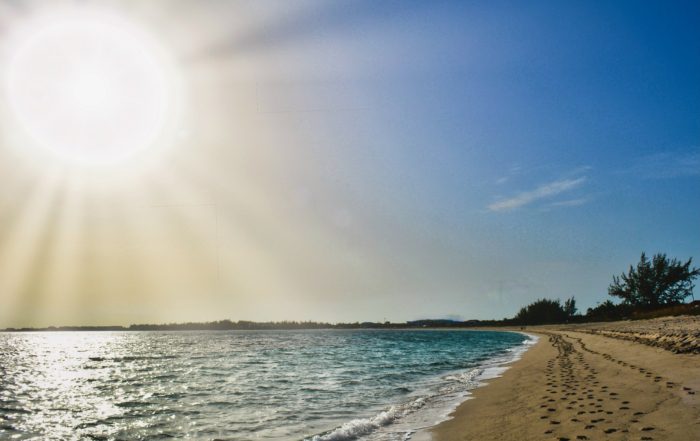 Sunlit beach scene with footprints in the sand, calm turquoise waves, and a large sun in the clear blue sky. The mood is serene and inviting.