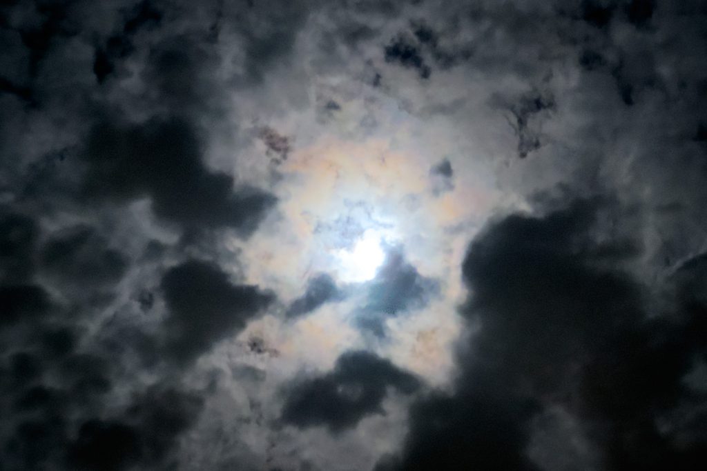 A bright moon partially obscured by dark clouds in a night sky. The light creates a mystical glow, highlighting scattered cloud patterns with hints of color.