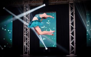 A dancer in a teal costume performs a high leap on stage, light beams highlighting her grace and skill. The backdrop displays glowing particles, enhancing the dynamic scene.