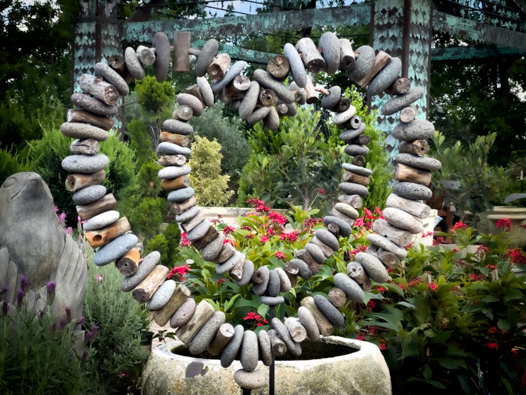 A heart-shaped sculpture made of stacked stones stands amid vibrant green foliage and red flowers. The scene is serene and earthy, with a nature-focused theme.