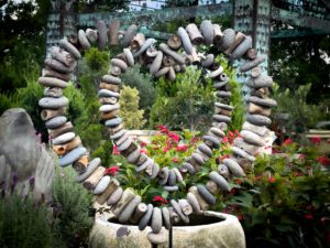 A heart-shaped sculpture made of stacked stones stands amid vibrant green foliage and red flowers. The scene is serene and earthy, with a nature-focused theme.