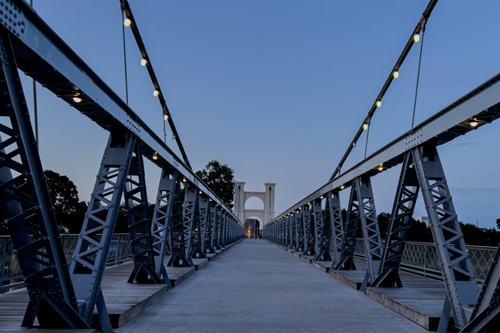 Steel suspension bridge at twilight with illuminated cables, creating a symmetrical view. The sky is a soft blue, evoking a calm ambiance.