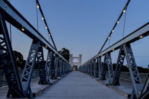 Steel suspension bridge at twilight with illuminated cables, creating a symmetrical view. The sky is a soft blue, evoking a calm ambiance.
