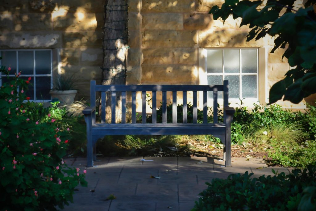 Wooden bench in a shaded garden area with flowering bushes, green plants, and a stone wall with two windows in the background.