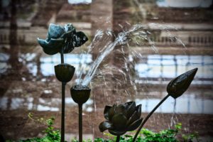 Decorative flower-shaped water fountain sprays water in arcs with blurred background reflections and greenery below.