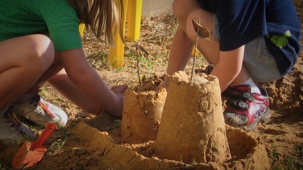Two children build sandcastles outdoors, using small spades. Focused and playful, they create towers adorned with twigs, embodying creativity and teamwork.