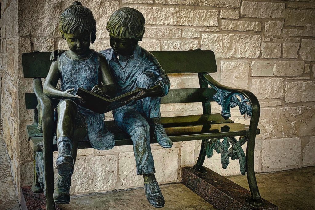 Bronze statue of two children sitting on a bench, reading a book together, set against a stone wall background.