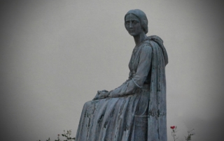 A serene statue of a seated woman in a flowing dress, gazing forward, with a single red rose nearby against a plain background, evoking introspection.