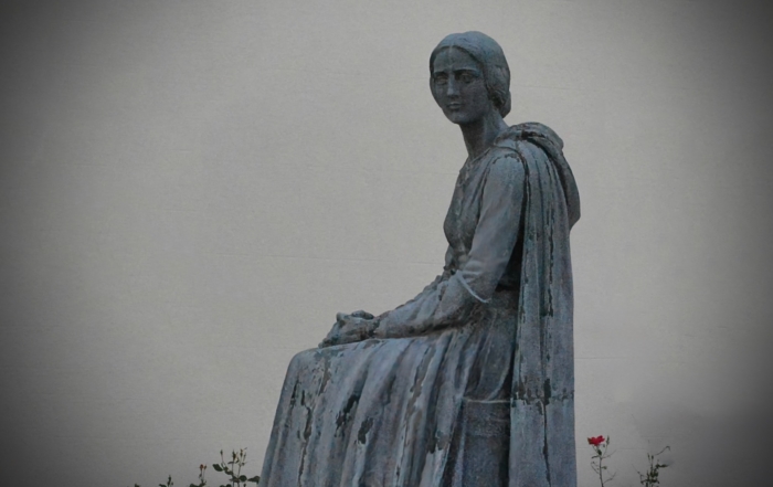 A serene statue of a seated woman in a flowing dress, gazing forward, with a single red rose nearby against a plain background, evoking introspection.