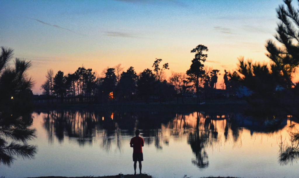 Person standing by a calm lake at sunset with silhouetted trees reflecting on the water.