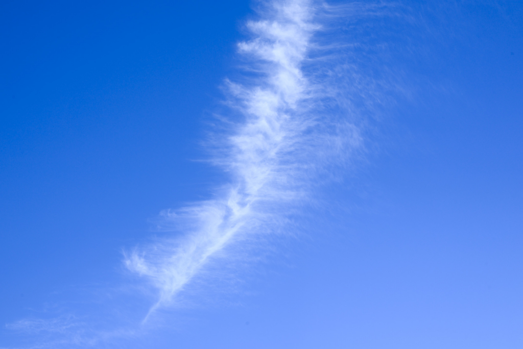 A wispy, white cloud stretches vertically against a vibrant blue sky, resembling a soft brushstroke. The scene conveys calm and tranquility.