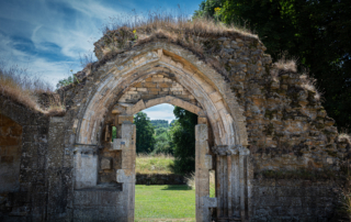 Ancient stone archway ruins with grass and trees visible through the opening under a partly cloudy blue sky.